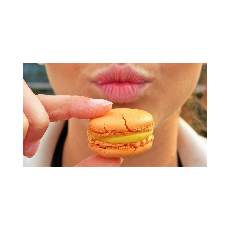 Macaron tenu entre deux doigts devant une bouche au second plan.
